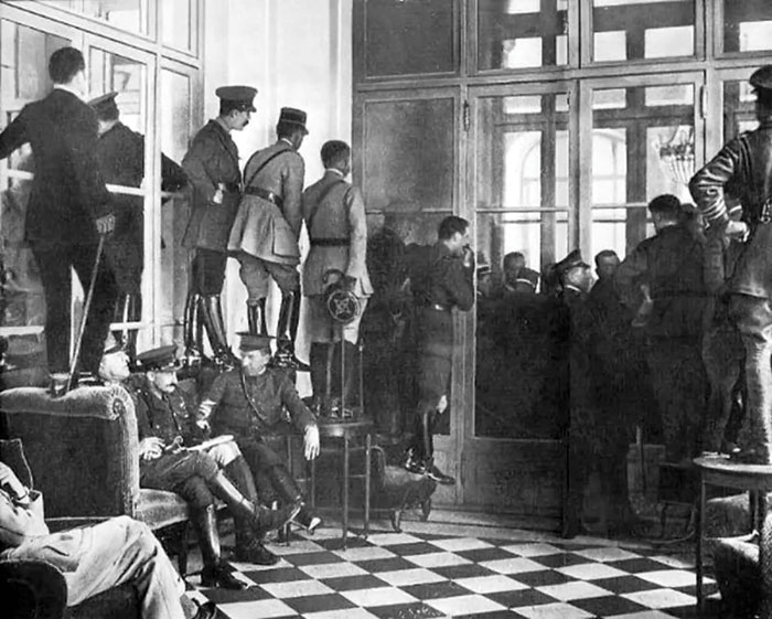 Black and white photograph showing a room of people, many stood on tables and chairs, peering through glass doors trying to catch a glimpse of the Treaty of Versailles being signed.