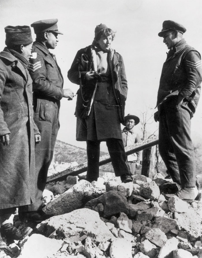 Black and white photograph of   Gellhorn in Italy during the Second World War - she stands on a pile of rubble in conversation with three other people.